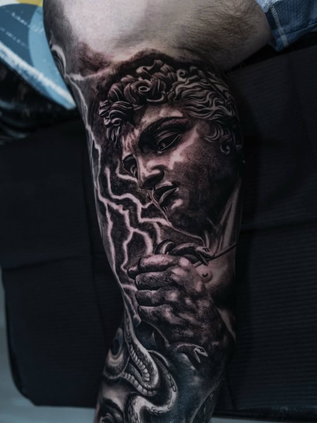 Classical male bust sculpture with curly hair and detailed facial features tattooed on thigh in monochrome greyscale.