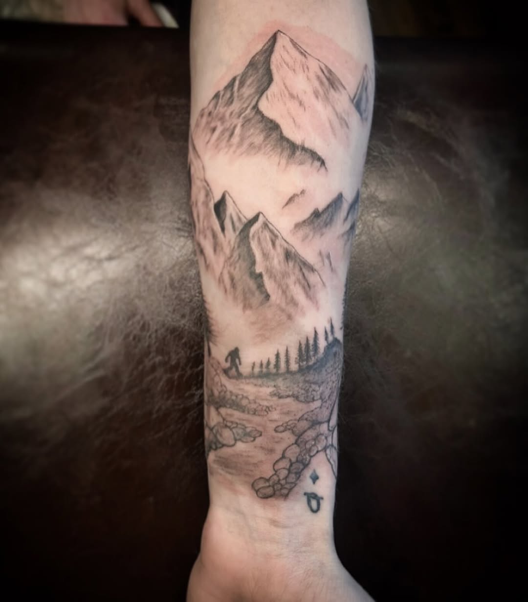 Black and grey mountain landscape tattoo on forearm featuring snow-capped peaks, evergreen trees, rocky terrain, and small decorative symbols.