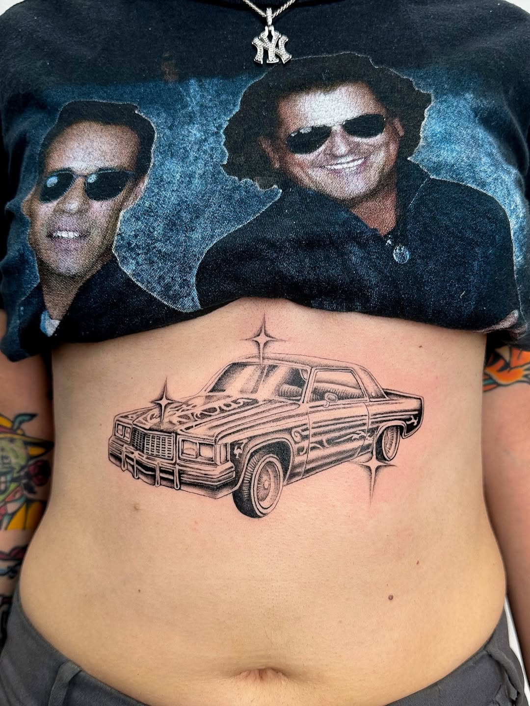 Black and grey lowrider car tattoo on chest with decorative cross above, fine detail shading