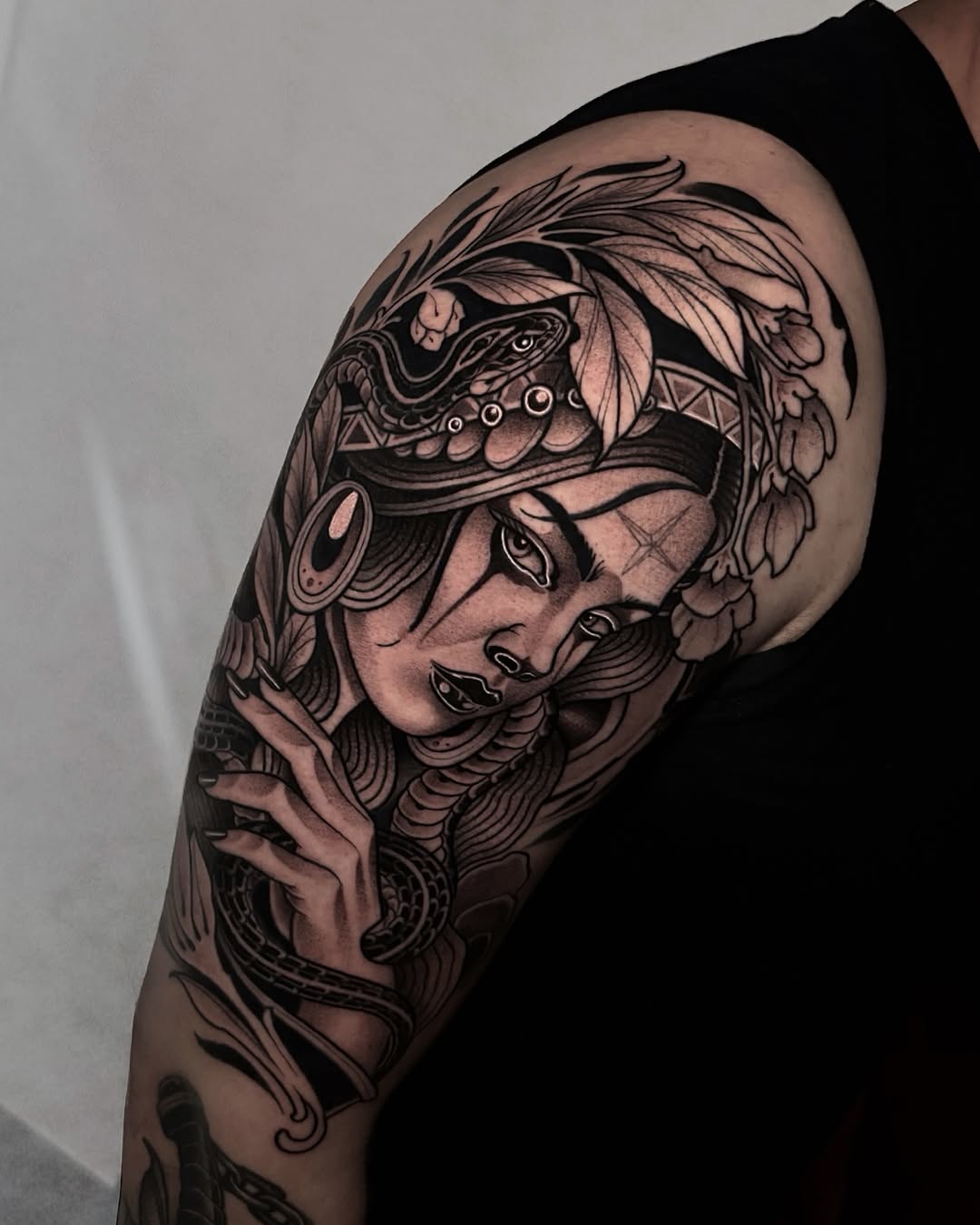 Black and grey portrait tattoo of a woman's face with ornate feathered headdress and decorative elements on upper arm.
