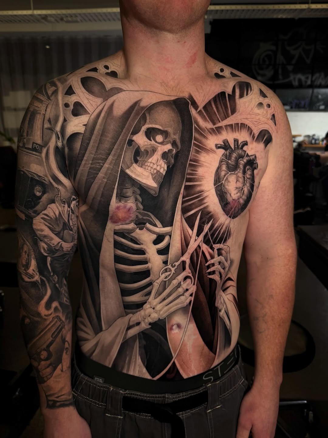 Full torso and arm tattoo featuring a hooded skeleton figure holding scissors, an anatomical heart, gothic architectural elements, and a crime-themed sleeve.