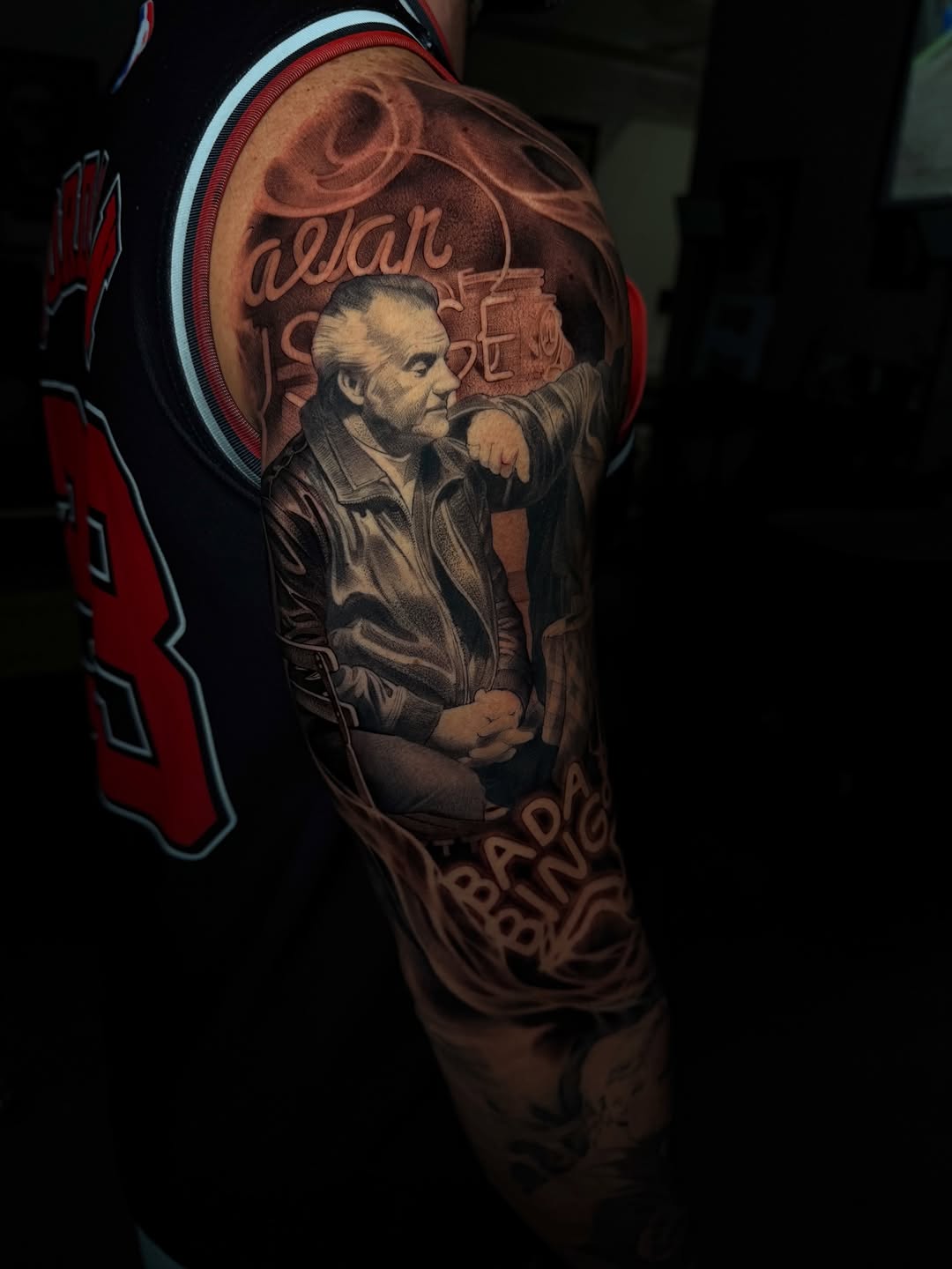 Black and grey arm tattoo depicting an elderly man in a jacket with pointing hand, lettering, and smoke swirl background.