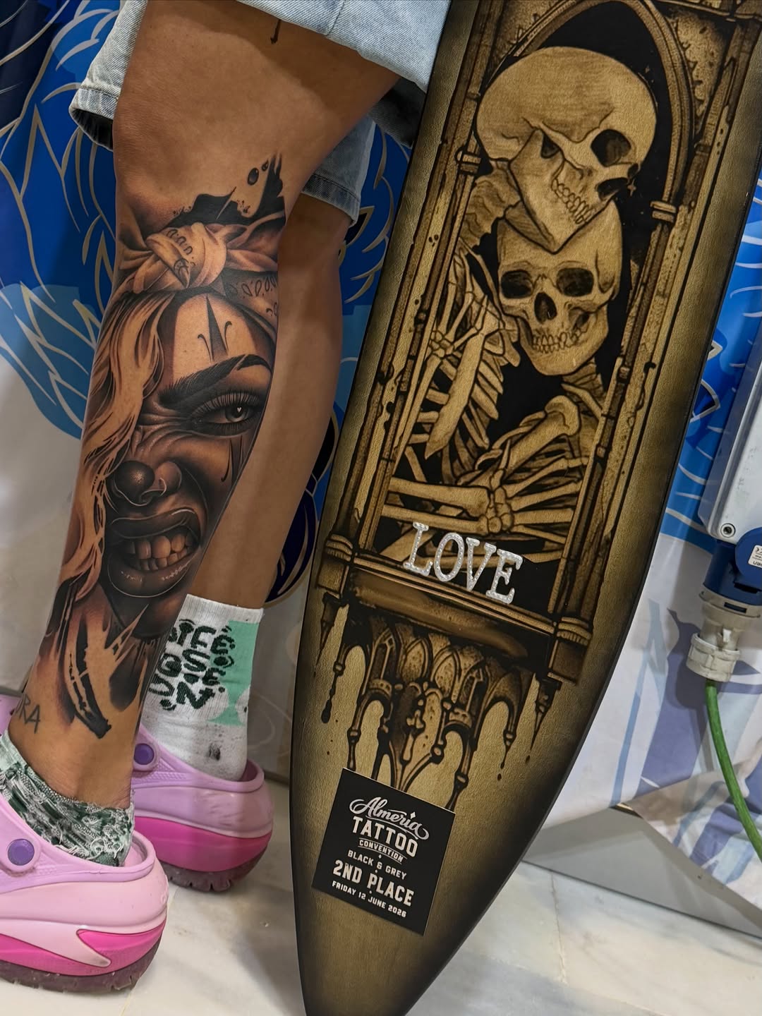 Black and grey portrait tattoo on leg featuring a woman's face with detailed shading and skull imagery above.