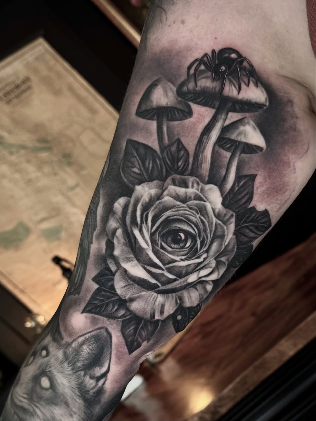 Black and grey tattoo on arm featuring a rose with an eye in the center, mushrooms above, and a spider, rendered in realistic shading.