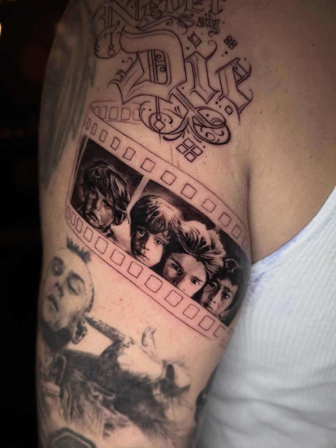 Black and grey portrait tattoo of four faces on upper arm with film strip border and decorative ornamental elements above.