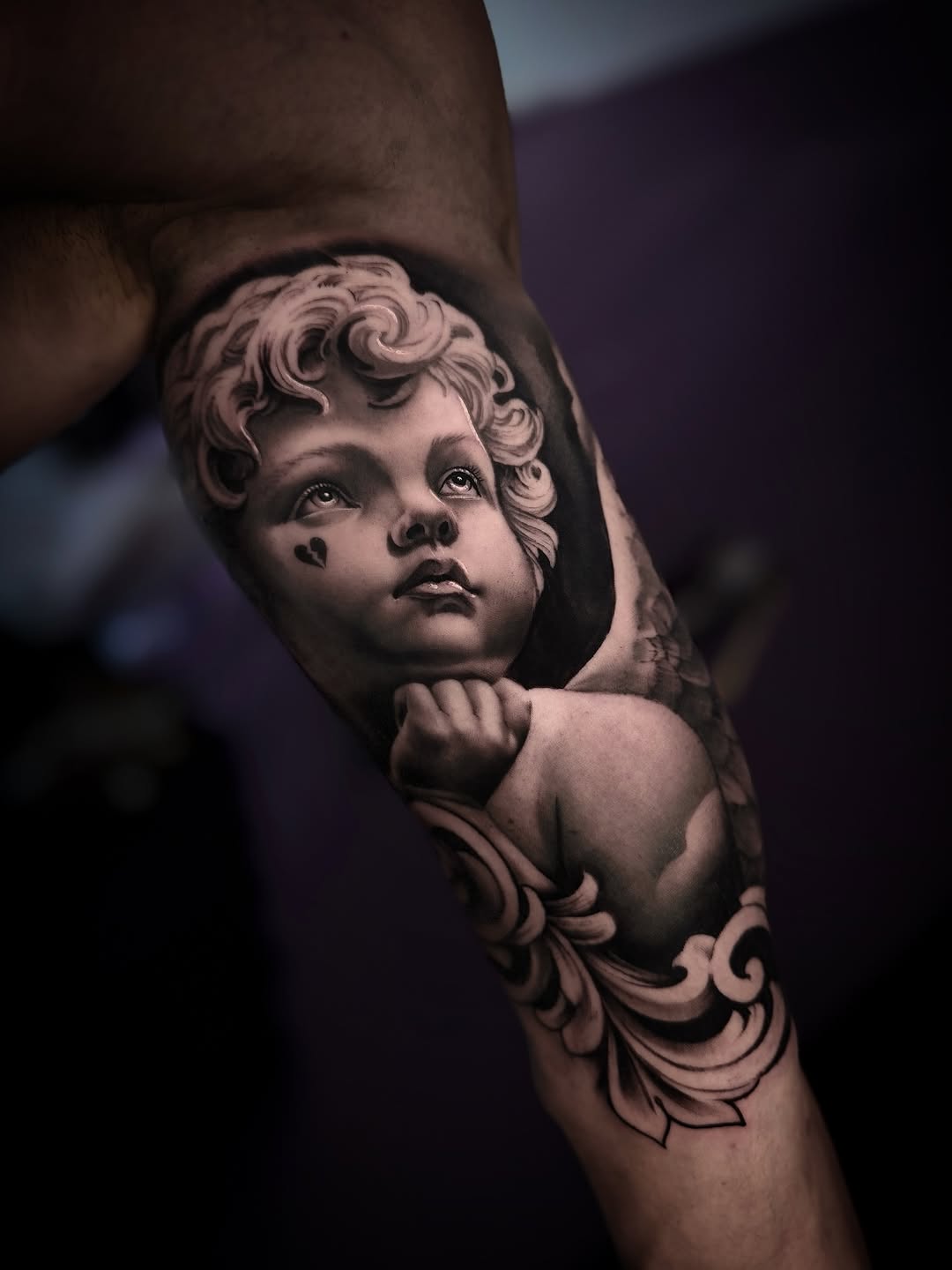 Cherub or baby face with curly hair, small heart on cheek, ornamental scrollwork below, rendered in black and grey on upper arm.