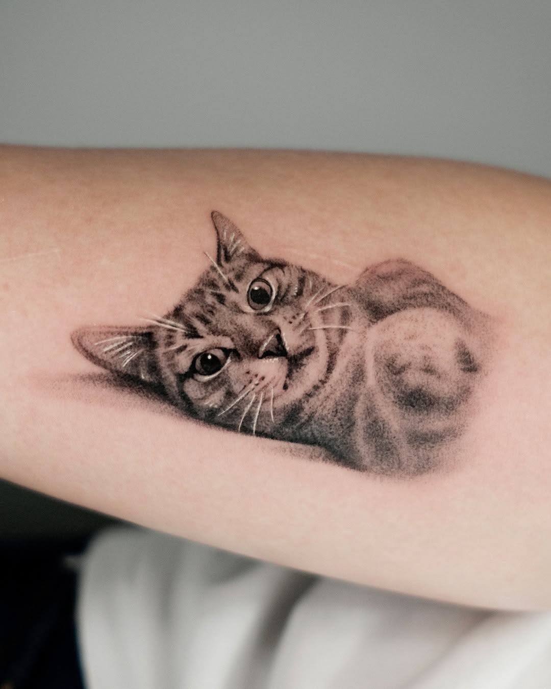 Black and grey tabby cat tattoo looking upward with detailed eyes and whiskers on forearm.