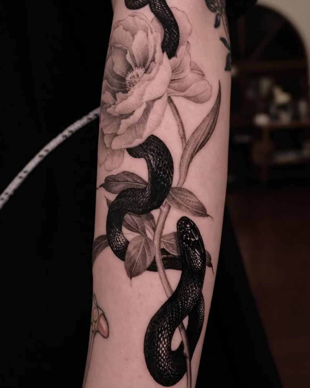 Black and grey tattoo of snakes intertwined with delicate flowers on the forearm, featuring detailed shading and botanical elements.