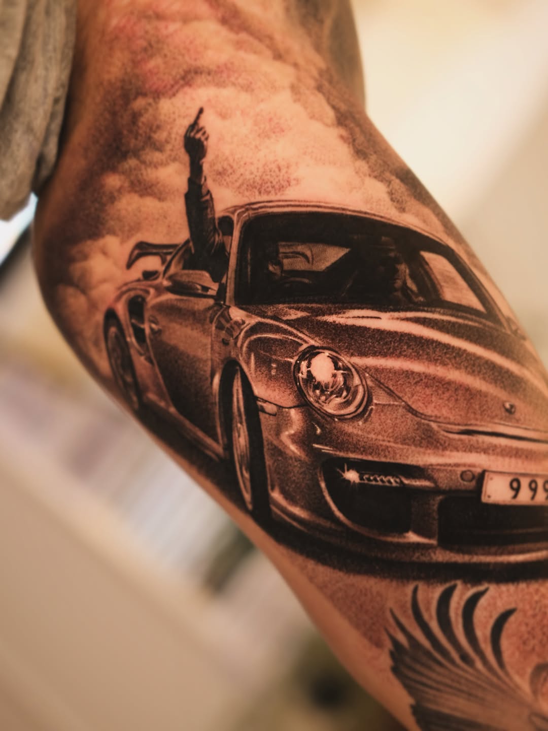 Black and grey realistic car tattoo on inner arm featuring a Porsche with open door and figure, detailed shading.