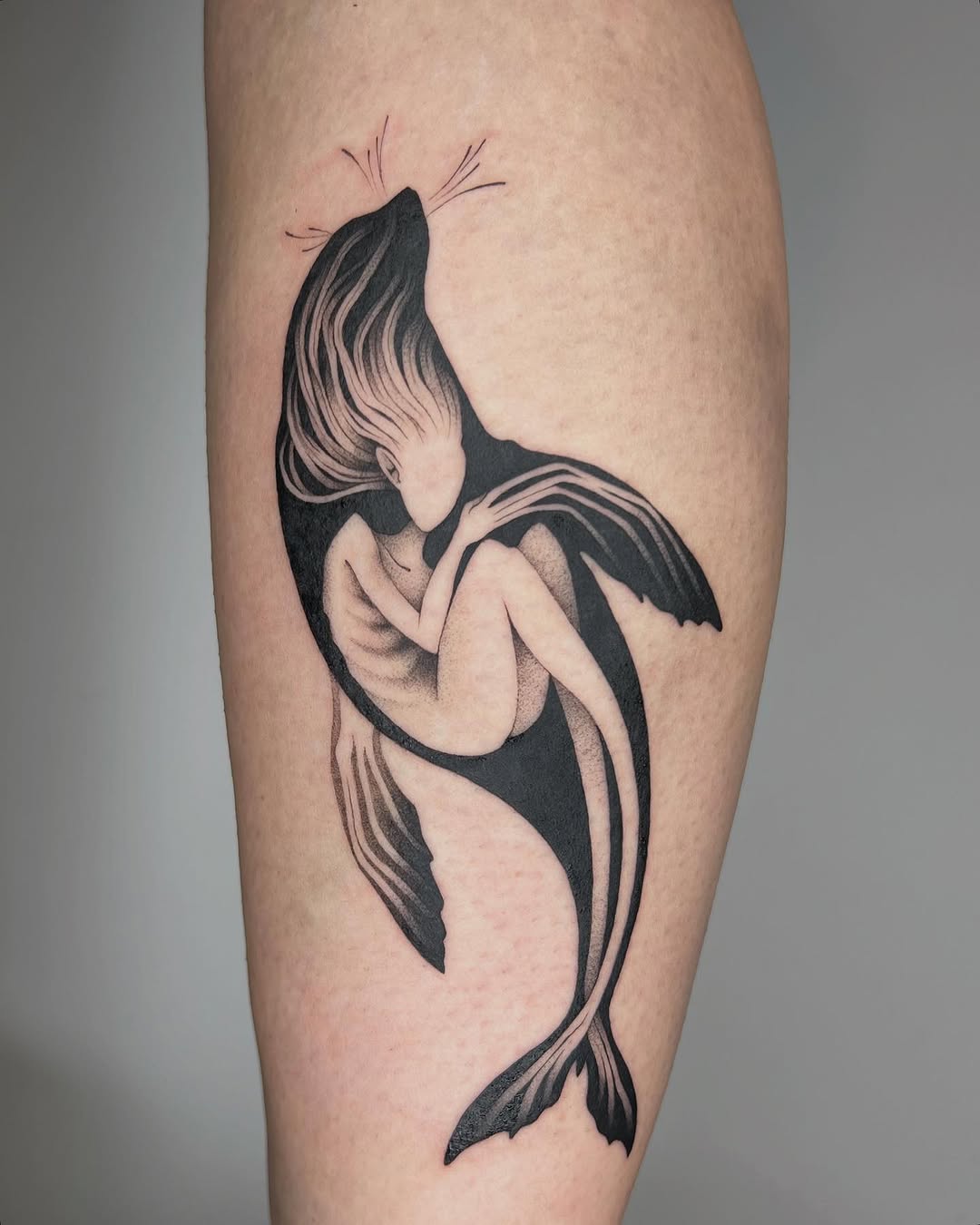 Black and grey mermaid tattoo on calf with flowing hair, detailed tail fins, and delicate whiskers.