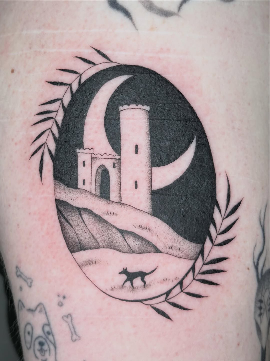 Black and grey tattoo of a crescent moon containing a desert fortress tower with a small fox below, framed by botanical leaf designs.