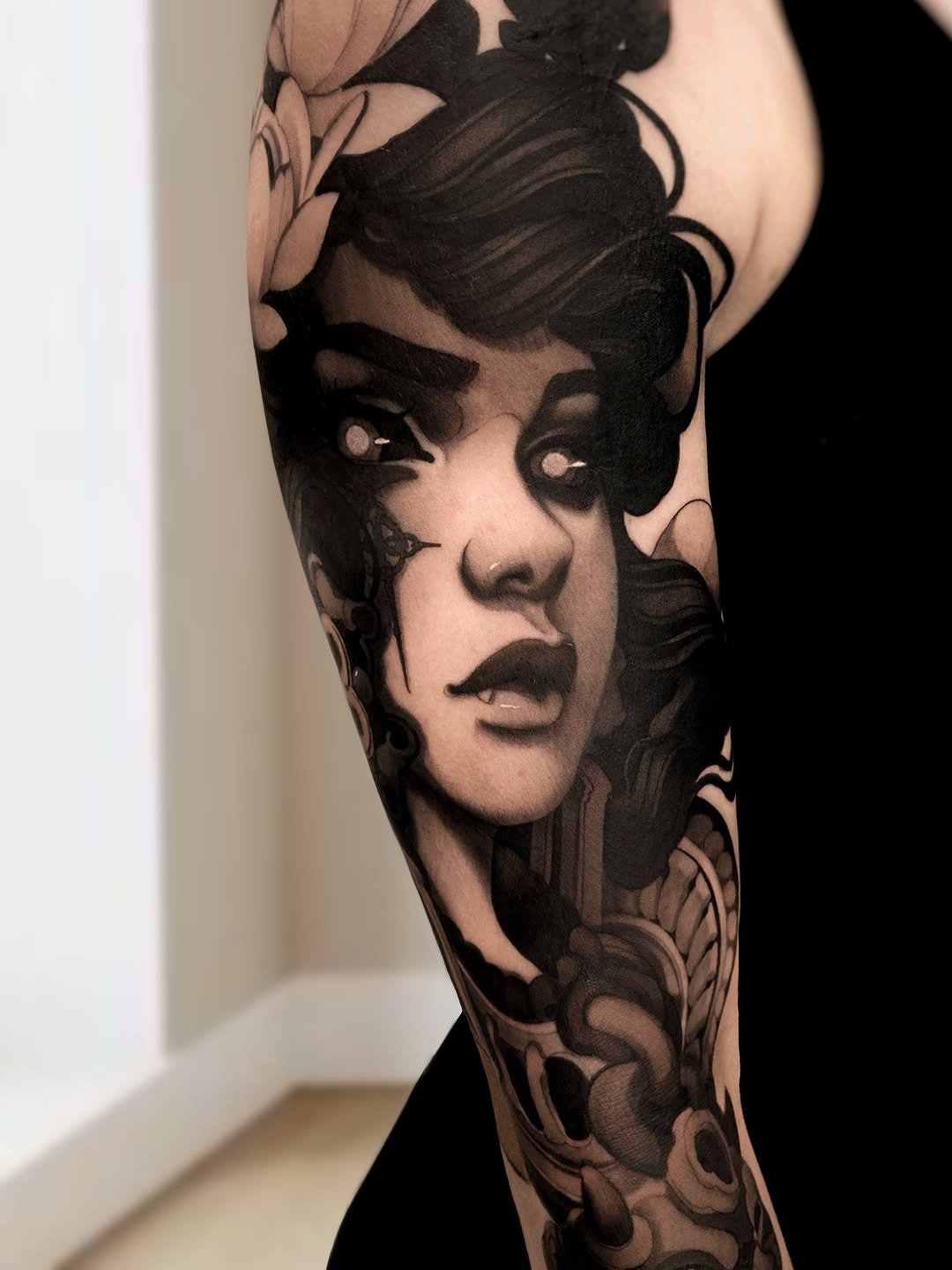Black and grey portrait tattoo of a woman's face with styled hair and decorative floral elements, on upper arm or shoulder.