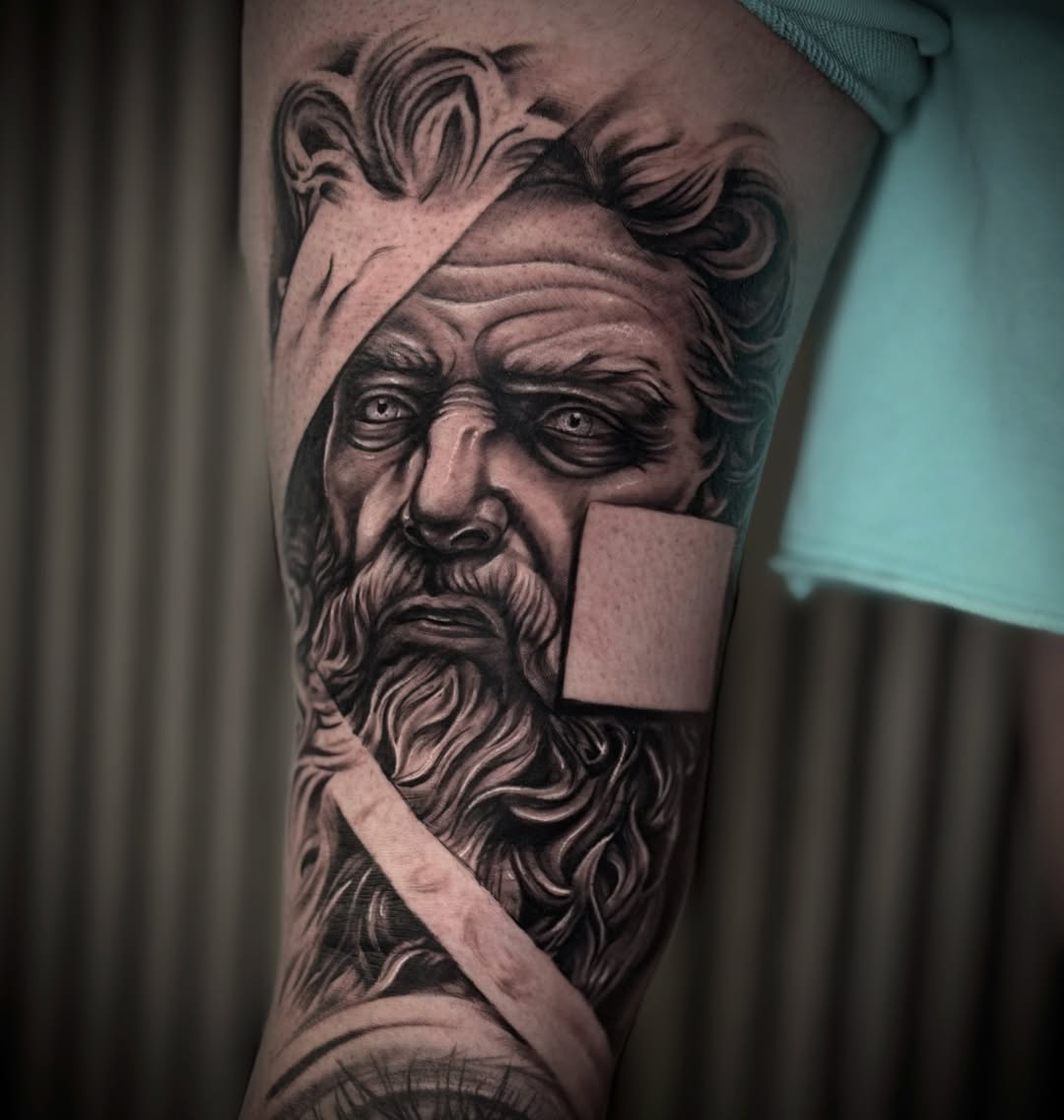 Black and grey portrait tattoo of a bearded man with curled hair on upper arm, with geometric rectangular shapes integrated into the design.