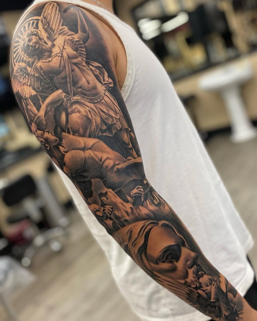 Black and grey full sleeve tattoo featuring St. Michael the Archangel with wings, armor, and demon, extending from shoulder to forearm.