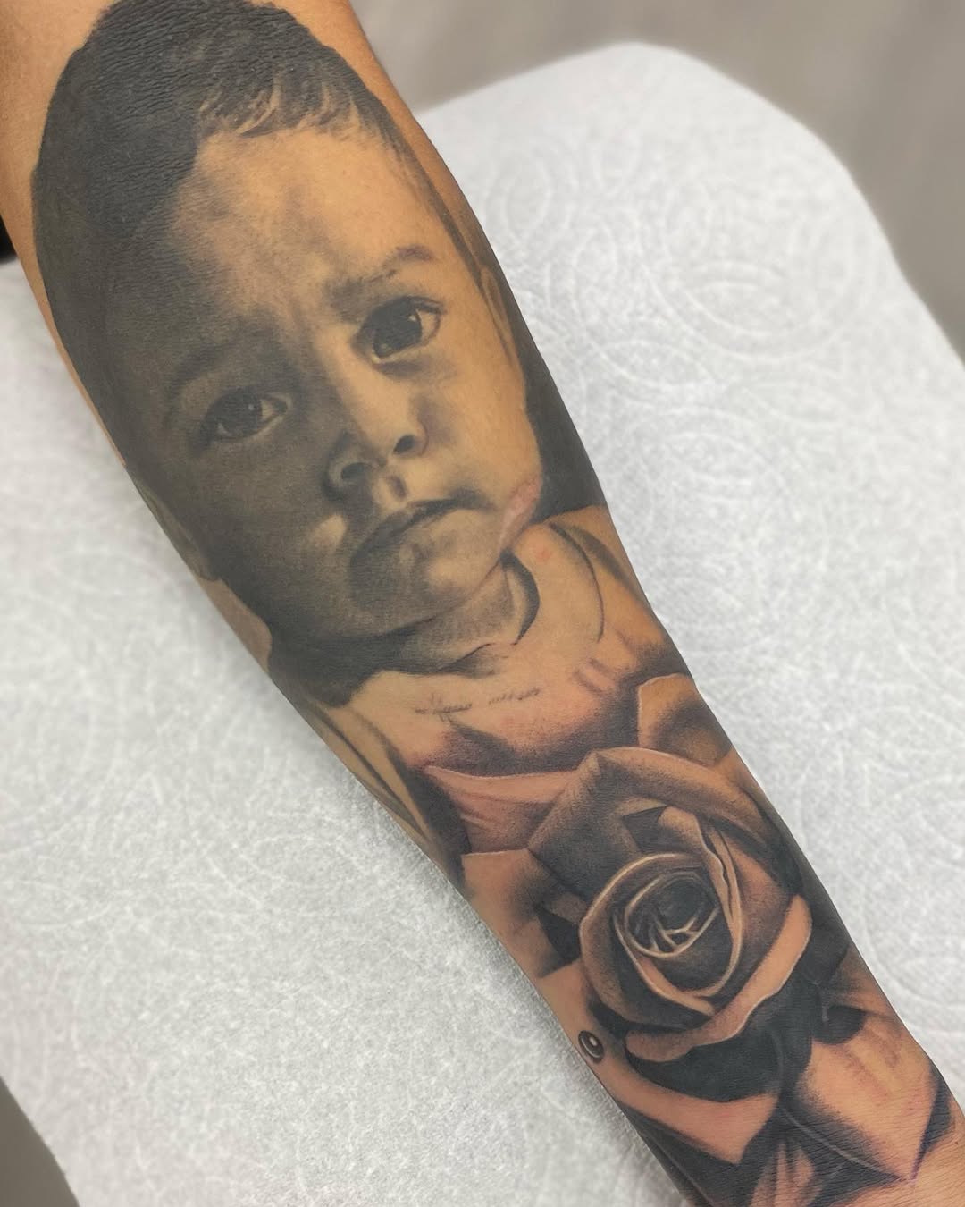 Black and grey portrait tattoo of a young child's face with a rose below on the forearm.