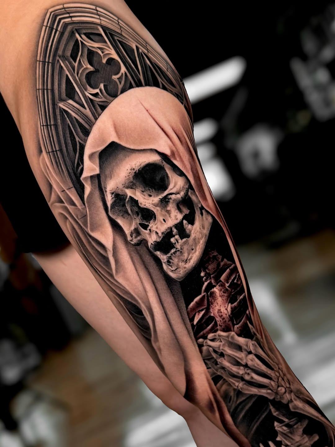 Reaper skull wearing hooded cloak with ornate geometric patterns on upper arm, rendered in black and grey with realistic shading.