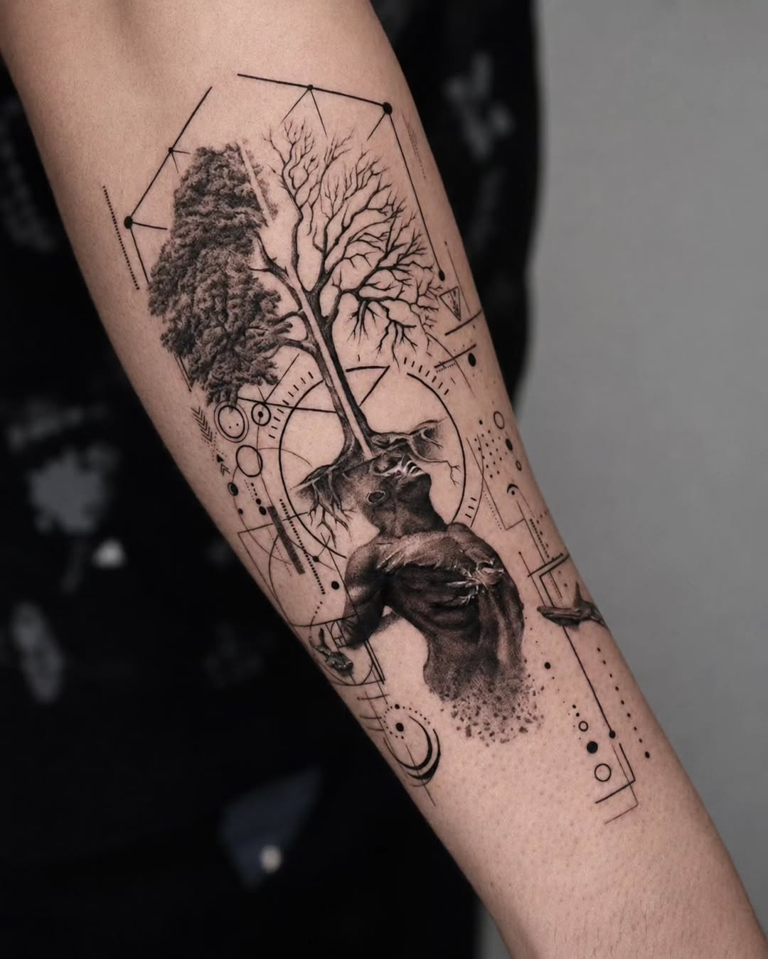 Black and grey arm tattoo featuring a tree with full foliage on one side, bare branches on the other, roots, geometric shapes, and a wolf head portrait.