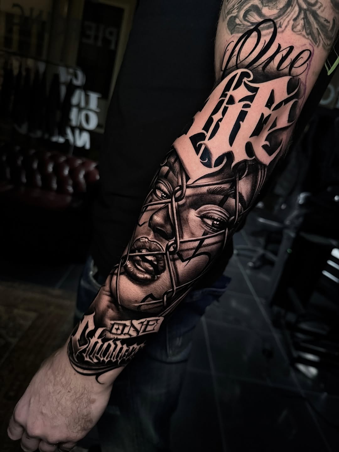 Black and grey sleeve tattoo featuring portrait faces with cursive text and decorative elements on forearm and upper arm.
