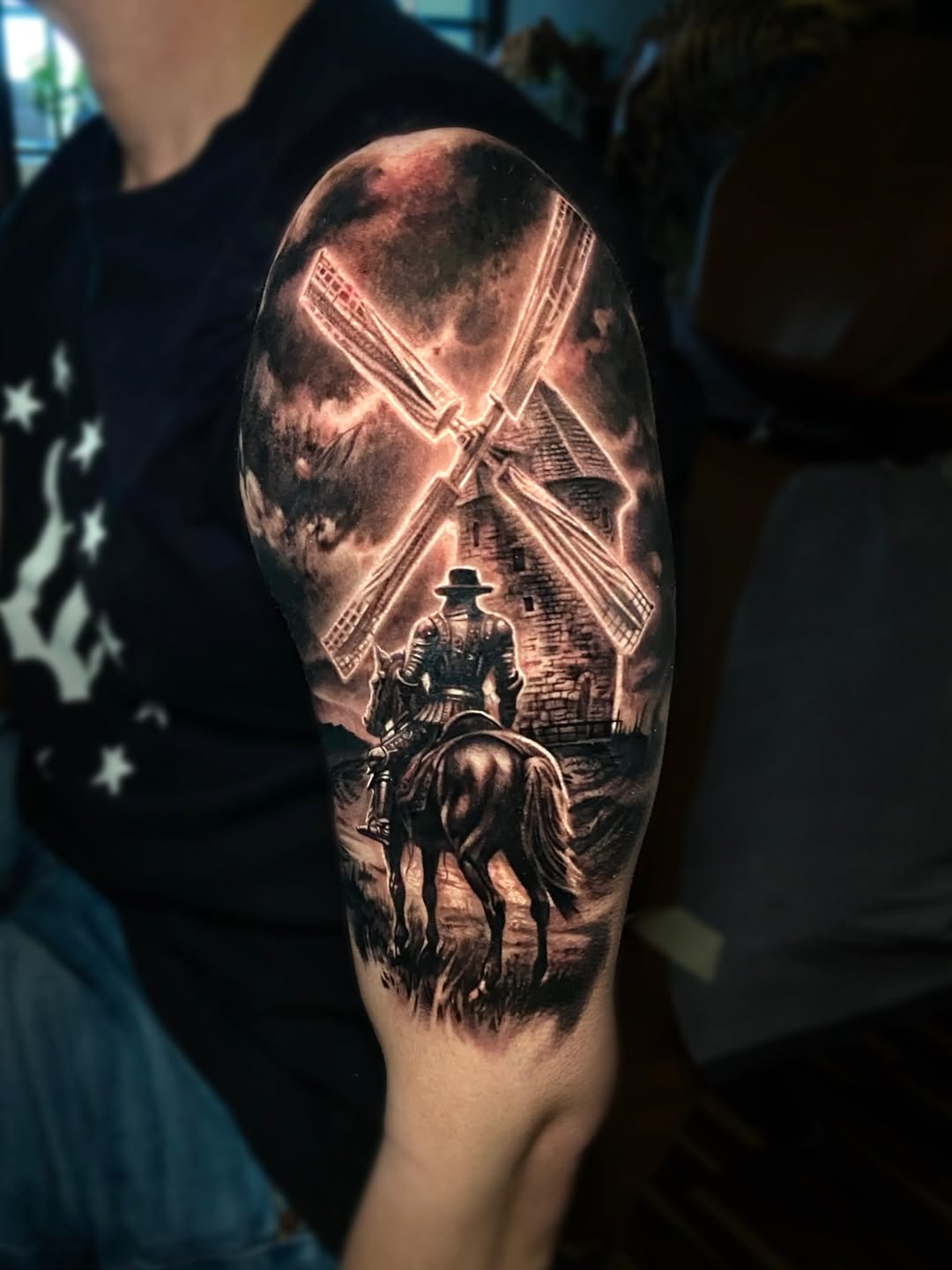 Upper arm tattoo depicting a rider on horseback approaching a windmill under a dramatic sky, rendered in black and grey shading.