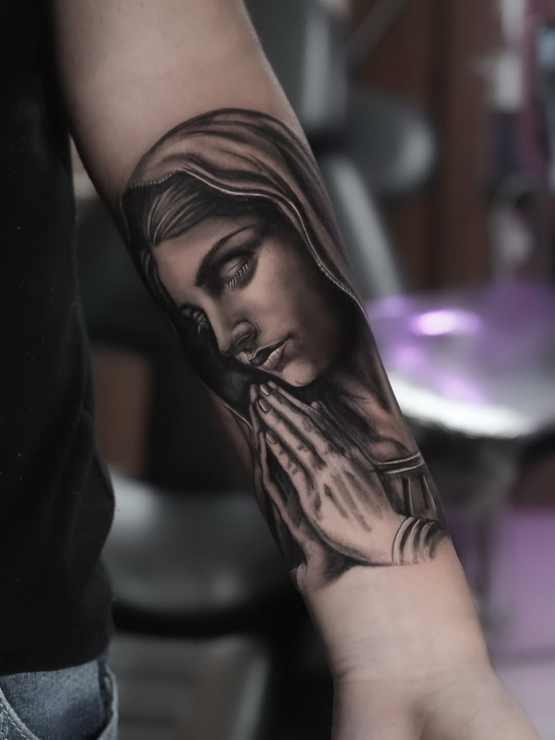 Black and grey portrait tattoo of a woman's face with long hair and praying hands on forearm.