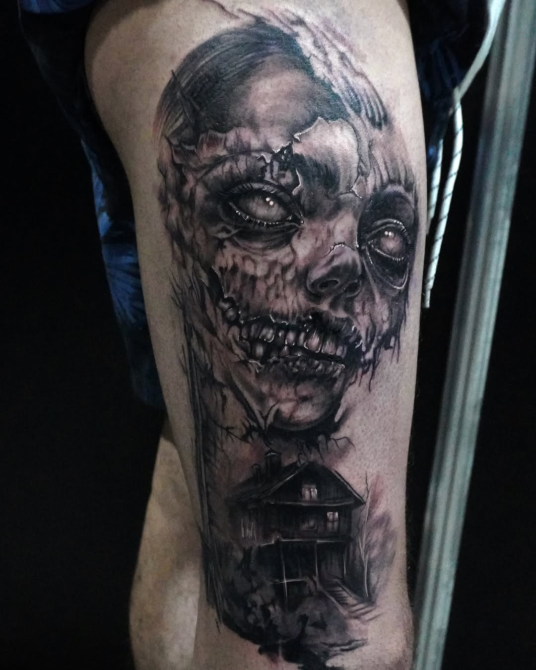 Black and grey portrait tattoo of a skull-faced figure with detailed shading on upper thigh, featuring a house below.