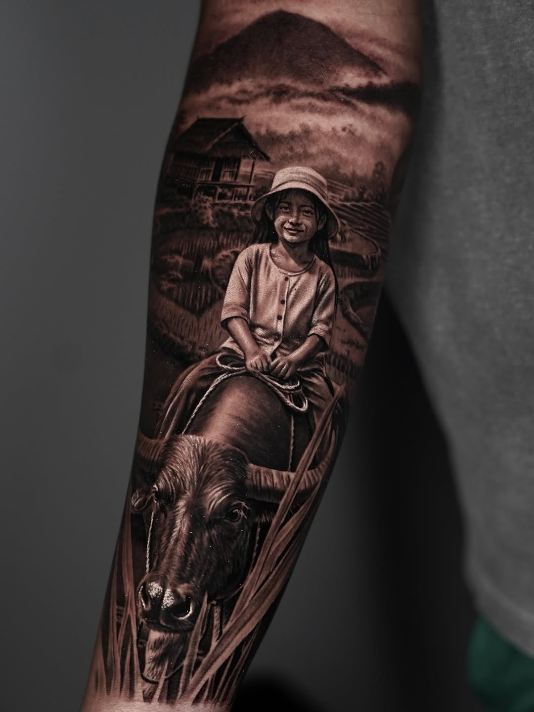 Child wearing a hat riding a water buffalo with a rural village and mountains in the background, on the forearm.