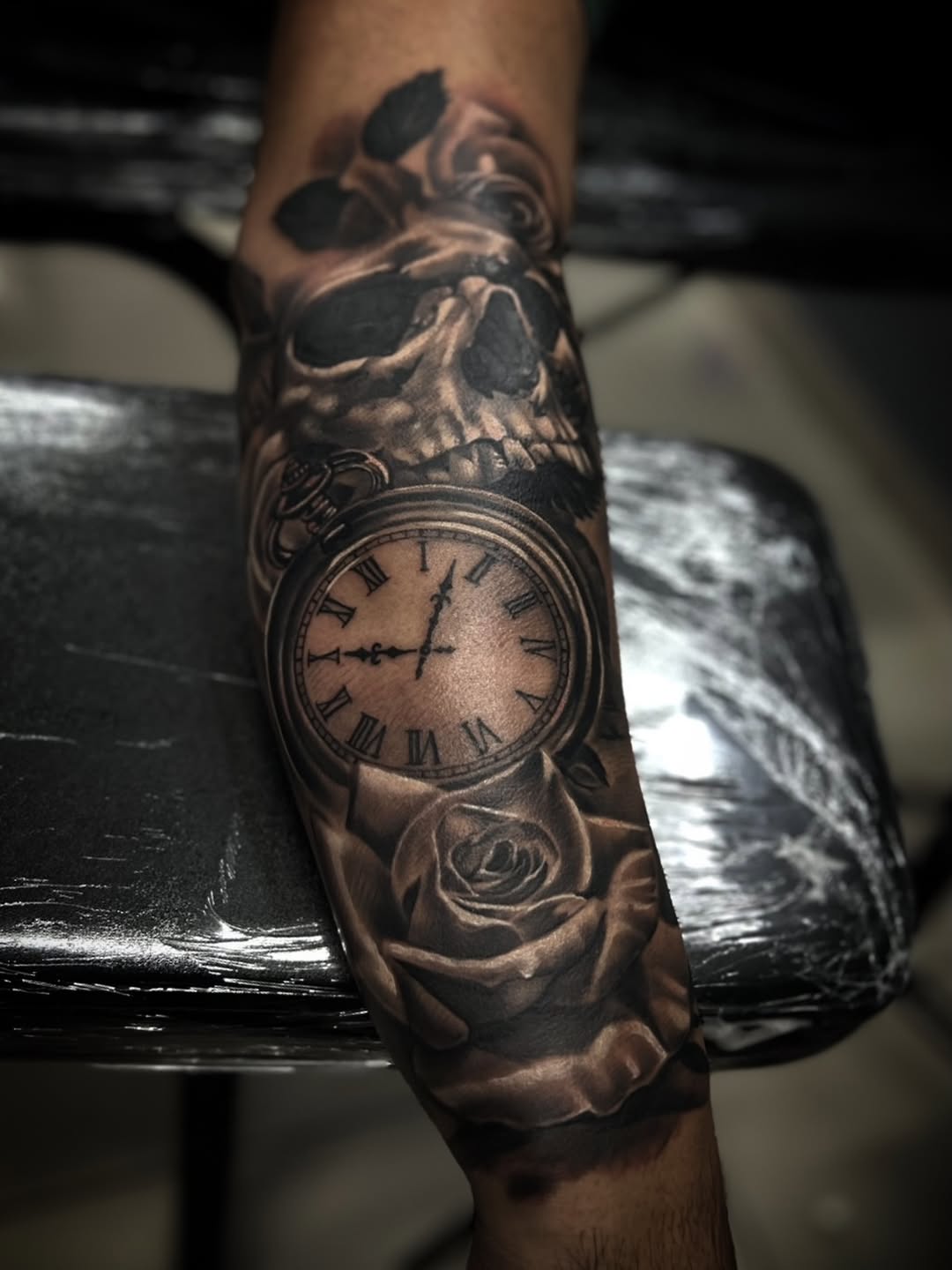 Monochromatic sleeve tattoo featuring a skull, pocket watch with Roman numerals, and rose on the forearm.