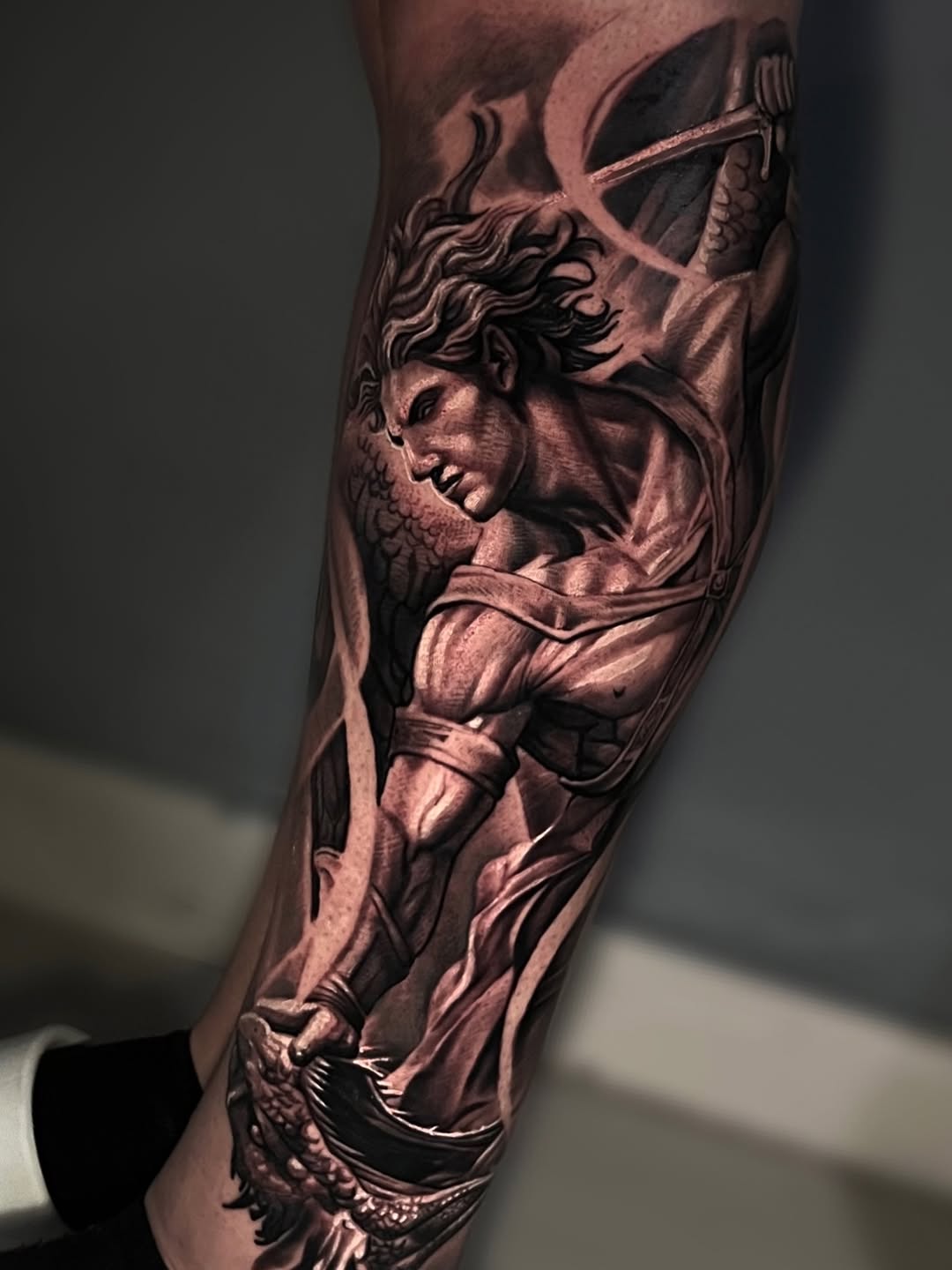 Classical male bust sculpture with curly hair and drapery, rendered in monochromatic black and grey on forearm.