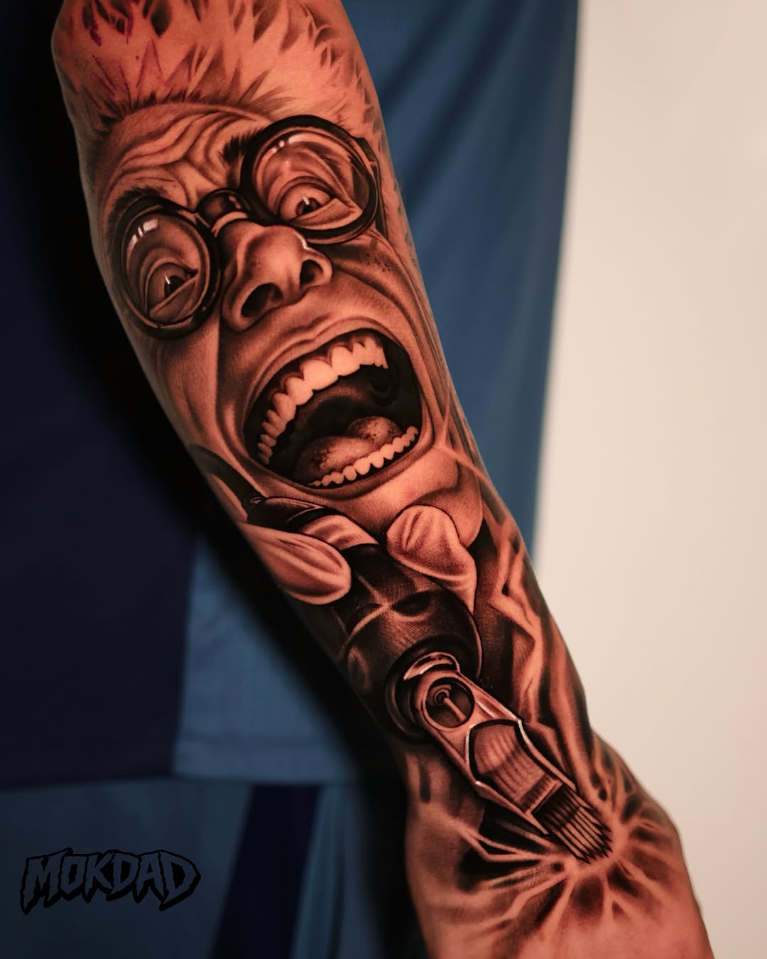 Black and grey realistic portrait tattoo of an expressive face with glasses and open mouth on forearm, with mechanical elements below.
