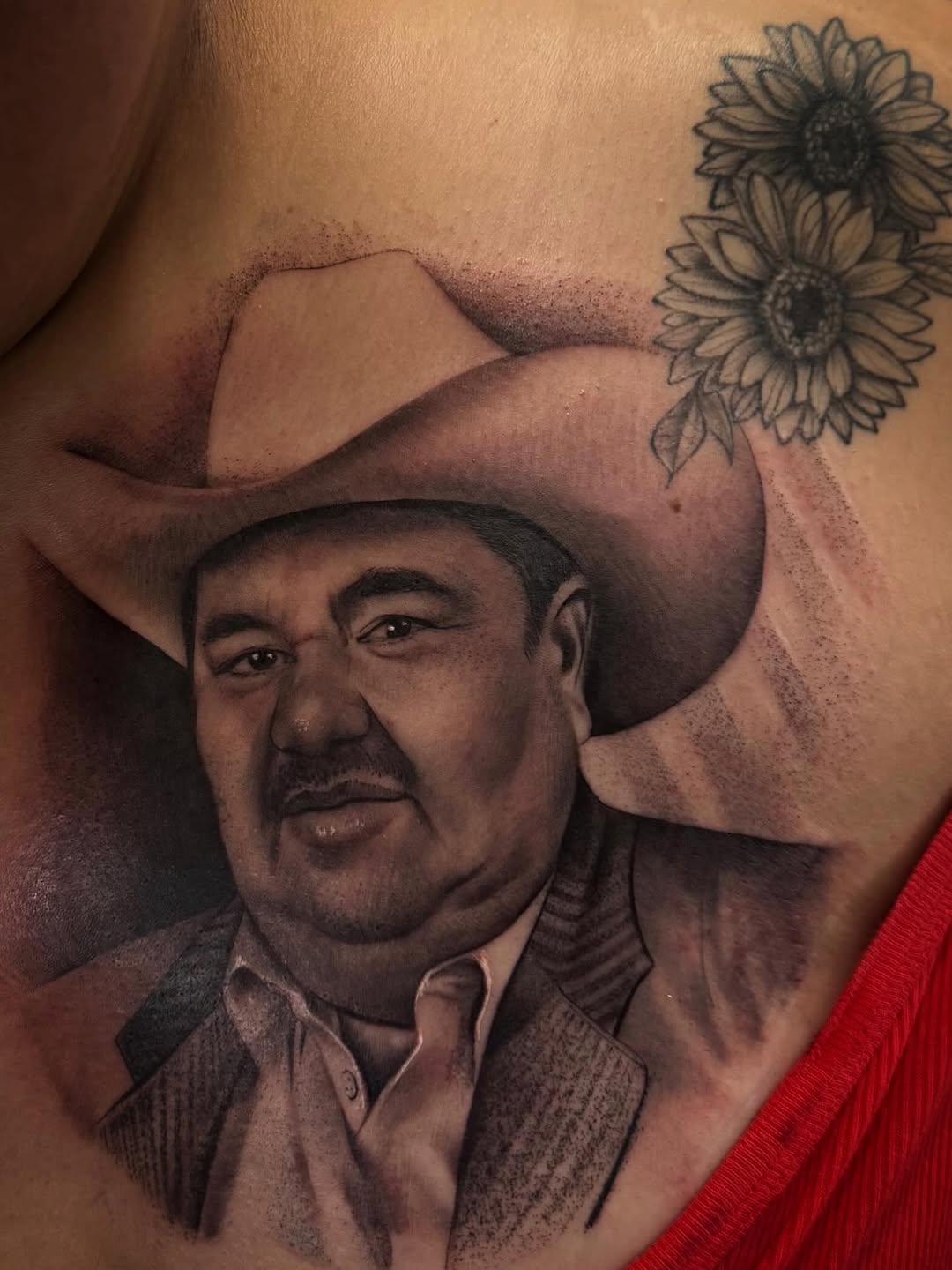 Black and grey portrait tattoo of a man wearing a cowboy hat with sunflowers above, placed on torso skin.