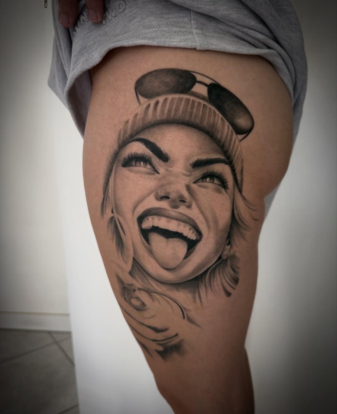 Black and grey portrait tattoo of a smiling woman wearing a beanie on the thigh, with detailed facial shading and decorative elements below.