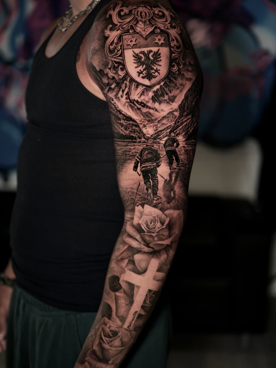 Full sleeve tattoo featuring a heraldic coat of arms with eagle, mountain landscape, two hikers, roses, and a cross in black and grey realism.