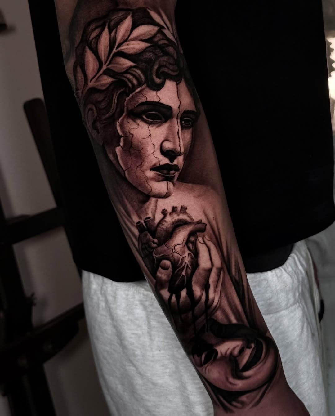 Black and grey portrait tattoo of a classical male face with laurel wreath crown and anatomical heart on forearm.