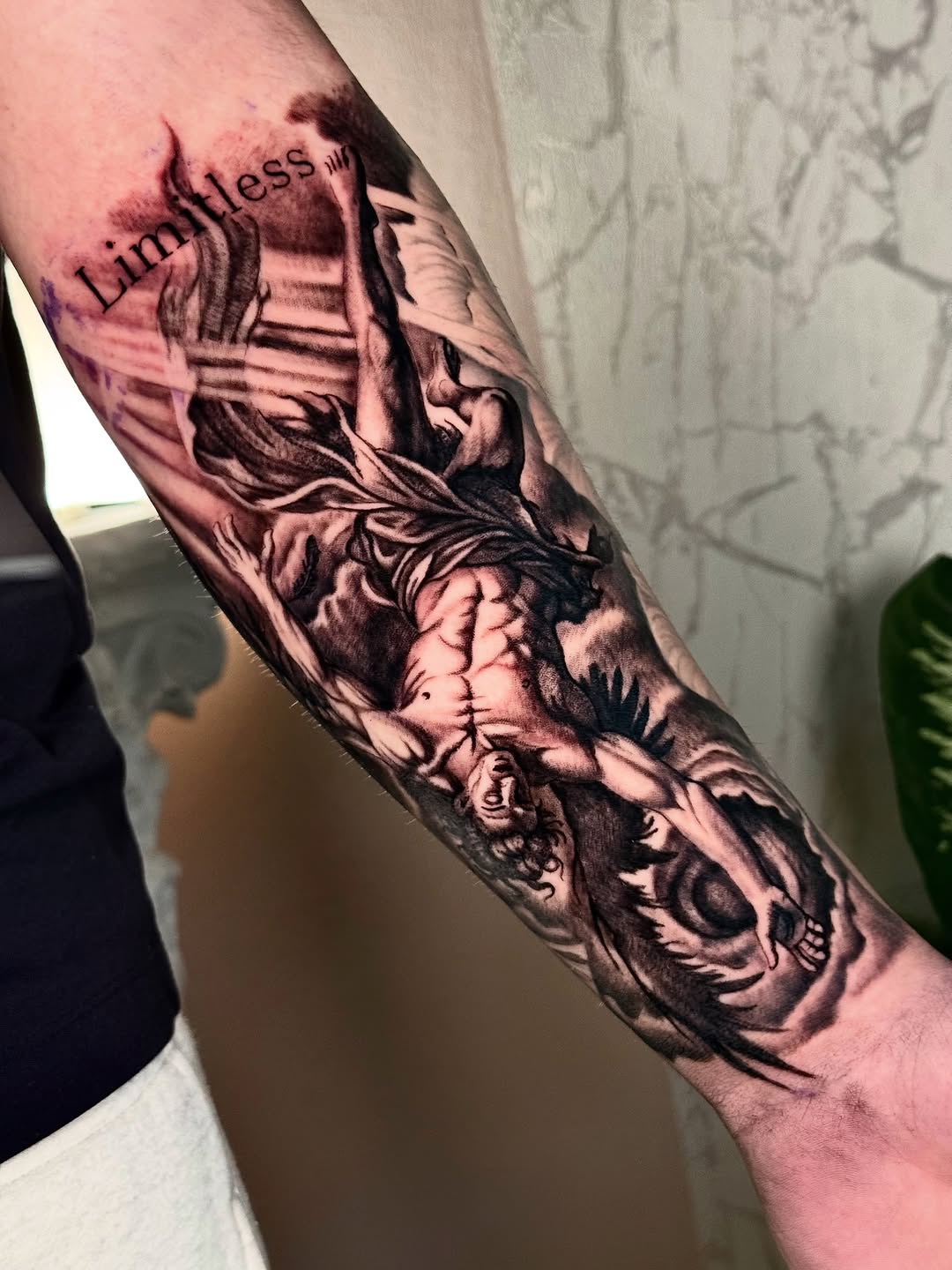 Black and grey realistic tattoo of a winged angel figure with 'Limitless' text on upper arm, featuring dramatic shading and anatomical detail.