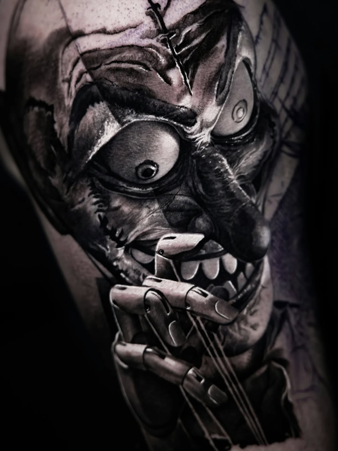Monochromatic demon or oni mask with fierce expression, sharp teeth, and ornamental hand gesture below, on upper arm.