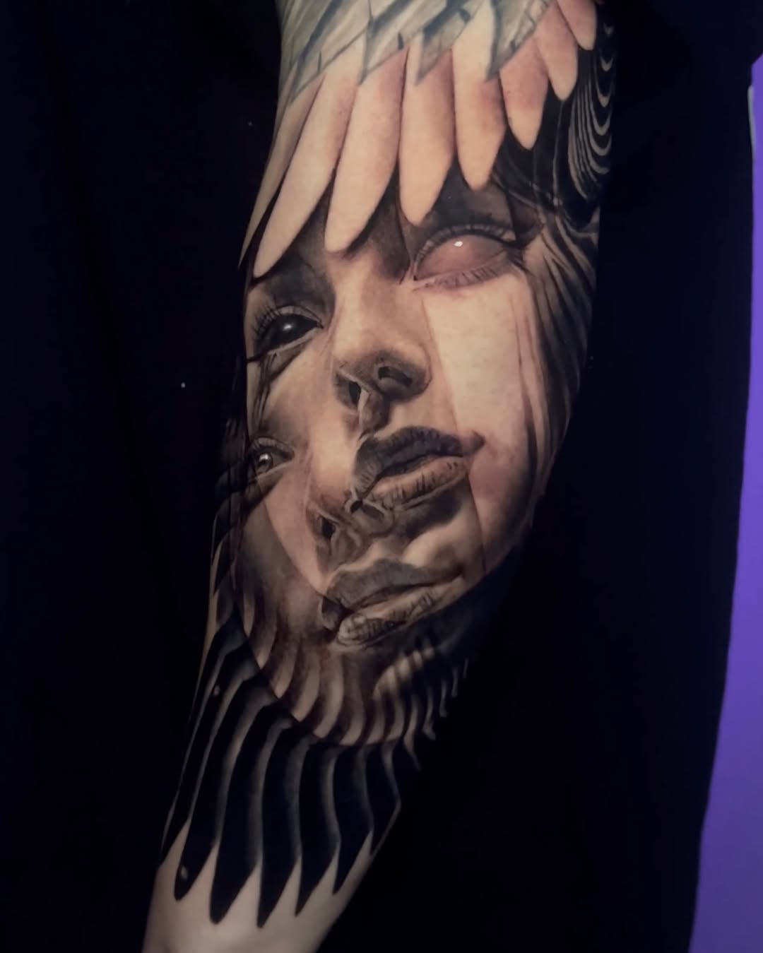 Black and grey portrait tattoo of a woman's face with bangs and feathered elements on upper arm, featuring soft shading and realism.