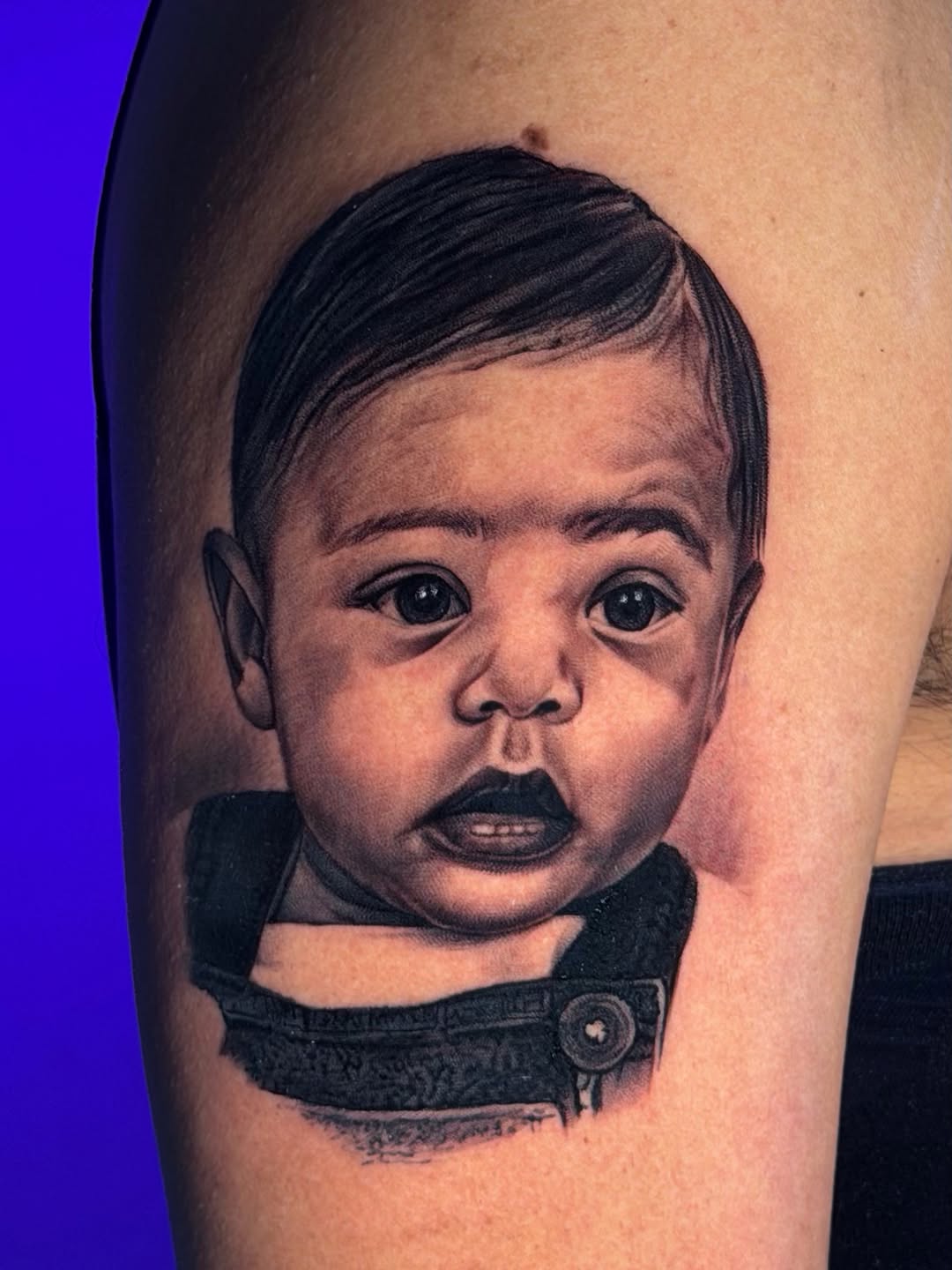 Black and grey portrait tattoo of a baby's face with surprised expression, wearing dark clothing, on arm.