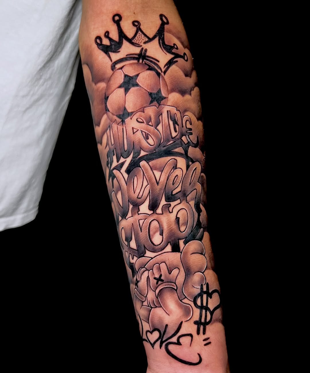 Black and grey tattoo on forearm featuring stacked graffiti-style text with crown, dollar sign, heart, and decorative elements.