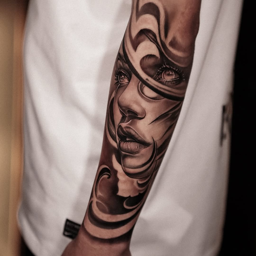 Monochromatic portrait of a woman's face with dramatic eye and lip detail, rendered in black and grey on the forearm.