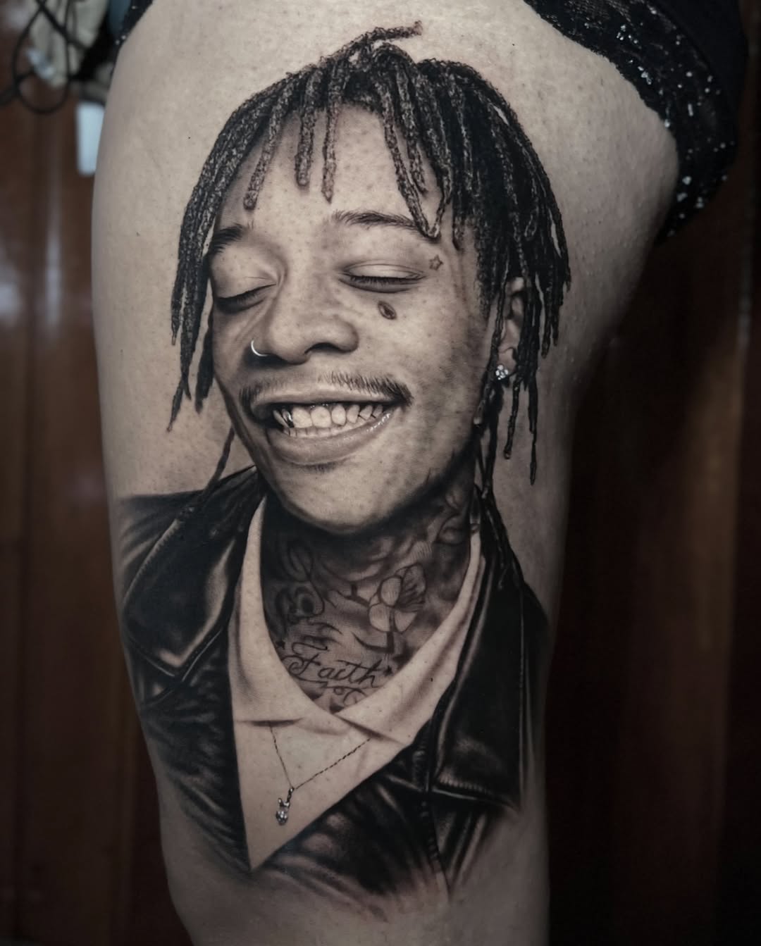 Black and grey portrait tattoo of a smiling person with braided hair and neck tattoos on upper arm.