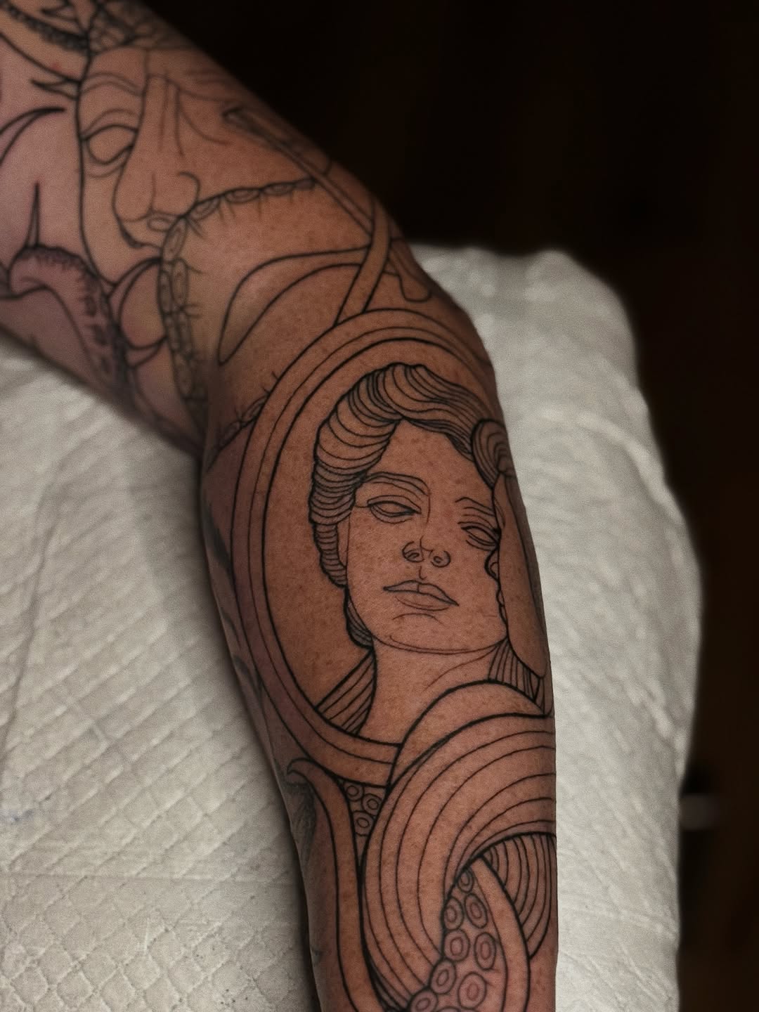 Black and grey portrait tattoo of a person's face with wavy hair on upper arm, surrounded by decorative line work and ornamental elements.