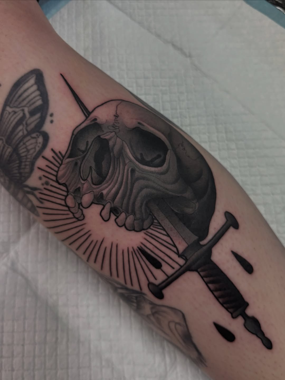Black and grey skull tattoo with wings, sword, and radiating line details on upper arm.