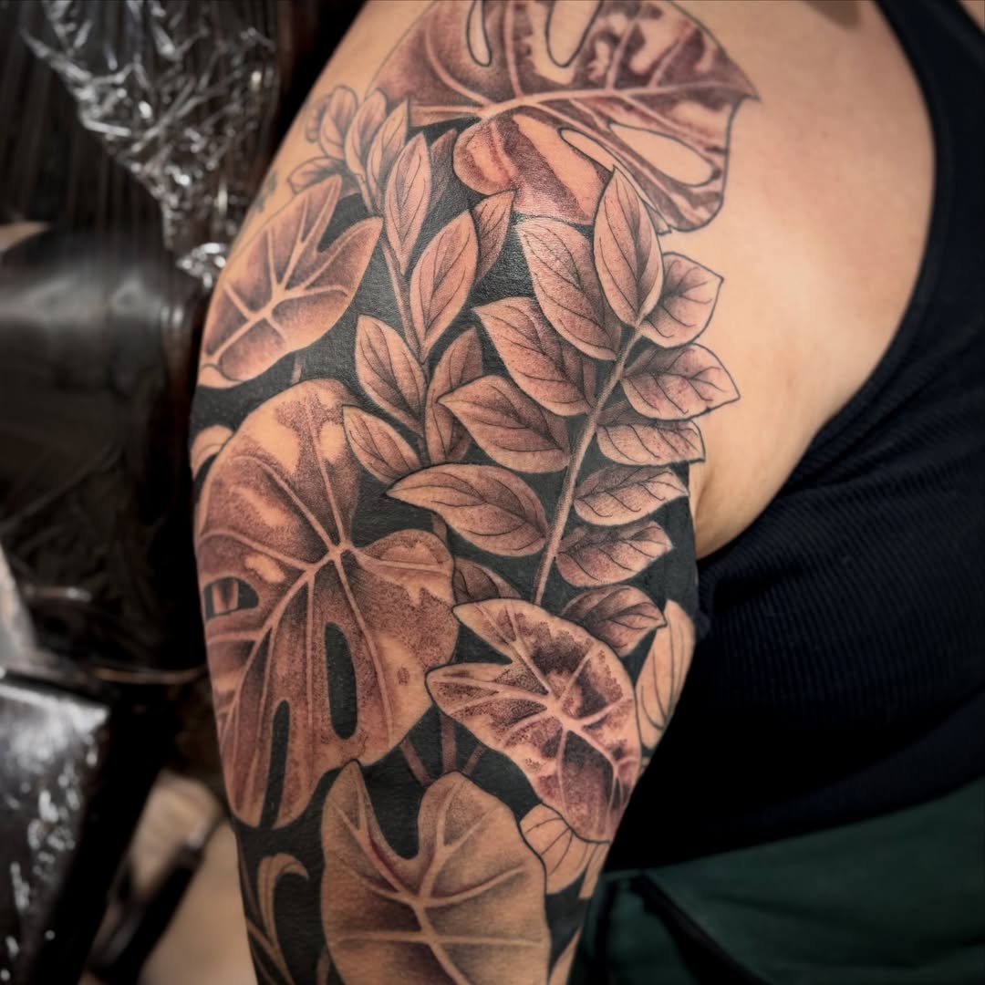 Black and grey botanical sleeve tattoo featuring detailed leaves and flowering plants with realistic shading on upper arm.