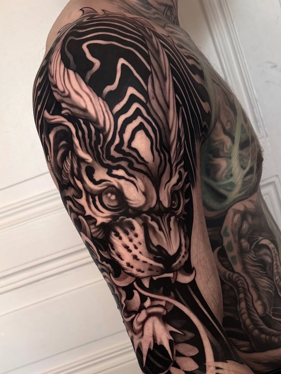Black and grey tiger head tattoo with intricate shading on upper arm, featuring detailed facial features and flowing mane.