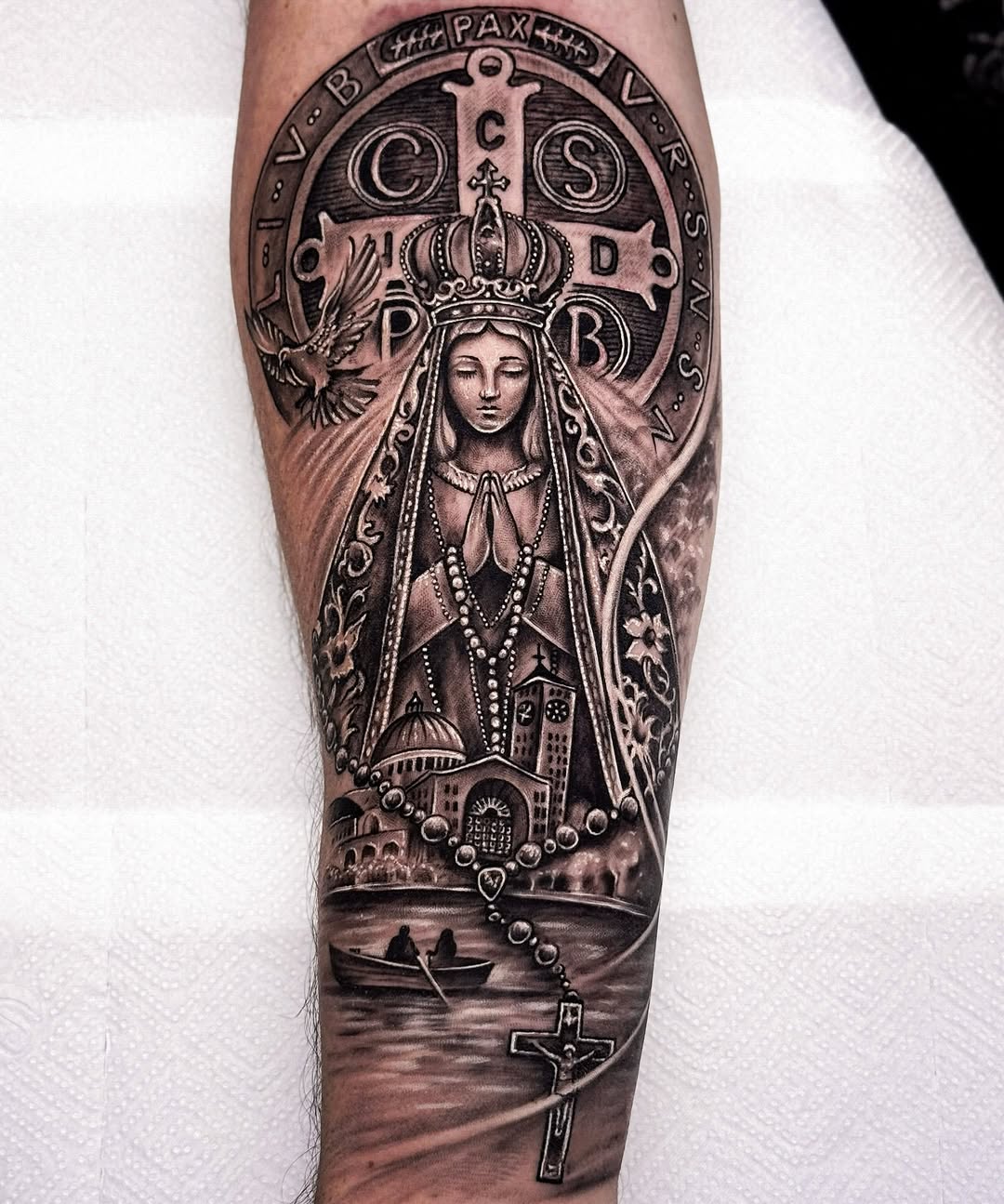 Religious sleeve tattoo featuring a crowned Virgin Mary figure with dove, ornate circular seal, rosary beads, church, boat, and cross in monochrome grey.