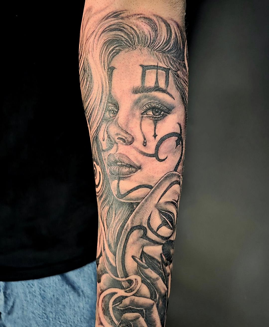 Black and grey portrait tattoo of a woman's face with wavy hair, tears, crescent moon, and decorative elements on forearm.