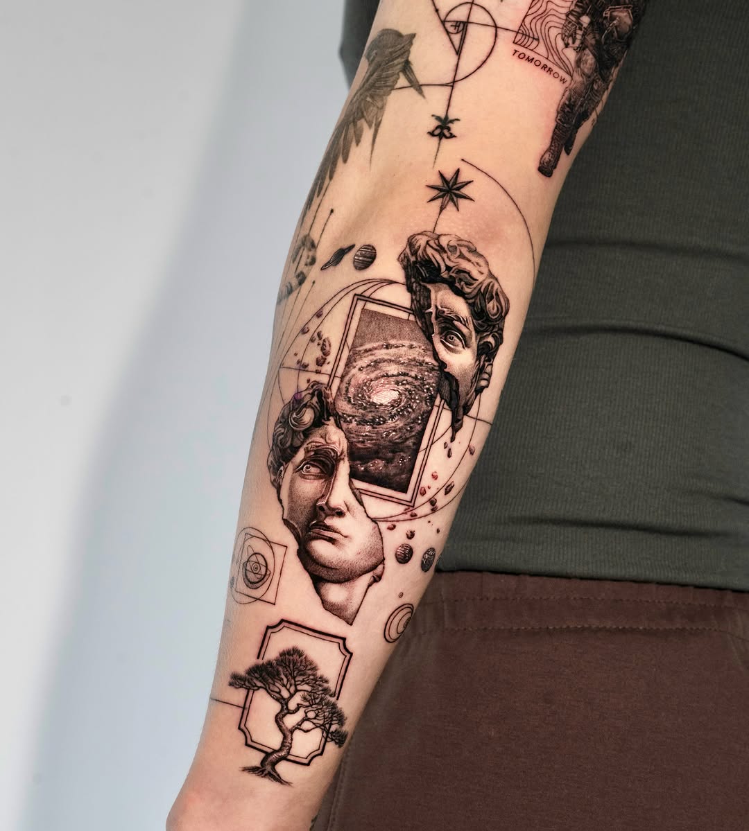 Black and grey surreal arm sleeve featuring classical busts, galaxies, stars, geometric shapes, and a bonsai tree in detailed realism.
