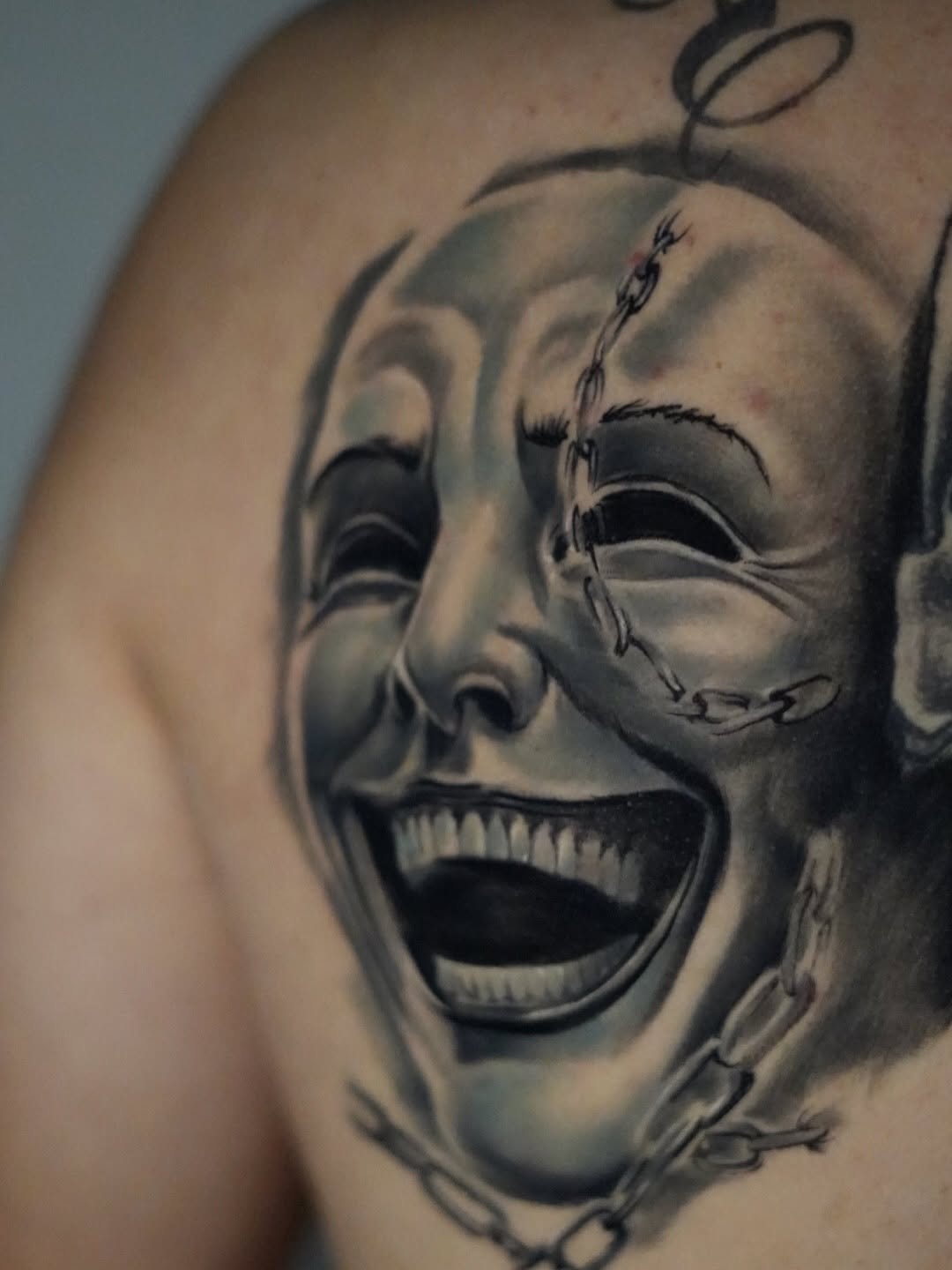 Black and grey realistic portrait tattoo of two theatrical masks showing contrasting emotions on upper arm, with stitched details.