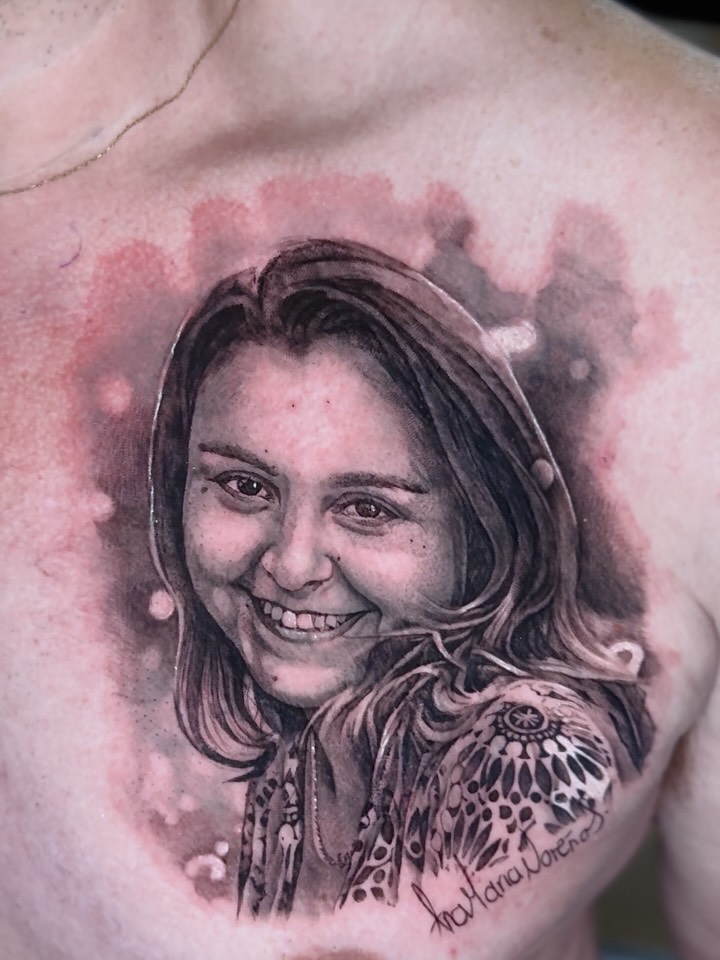 Black and grey chest tattoo of a smiling woman's portrait with a decorative floral shoulder piece and cursive name signature below.