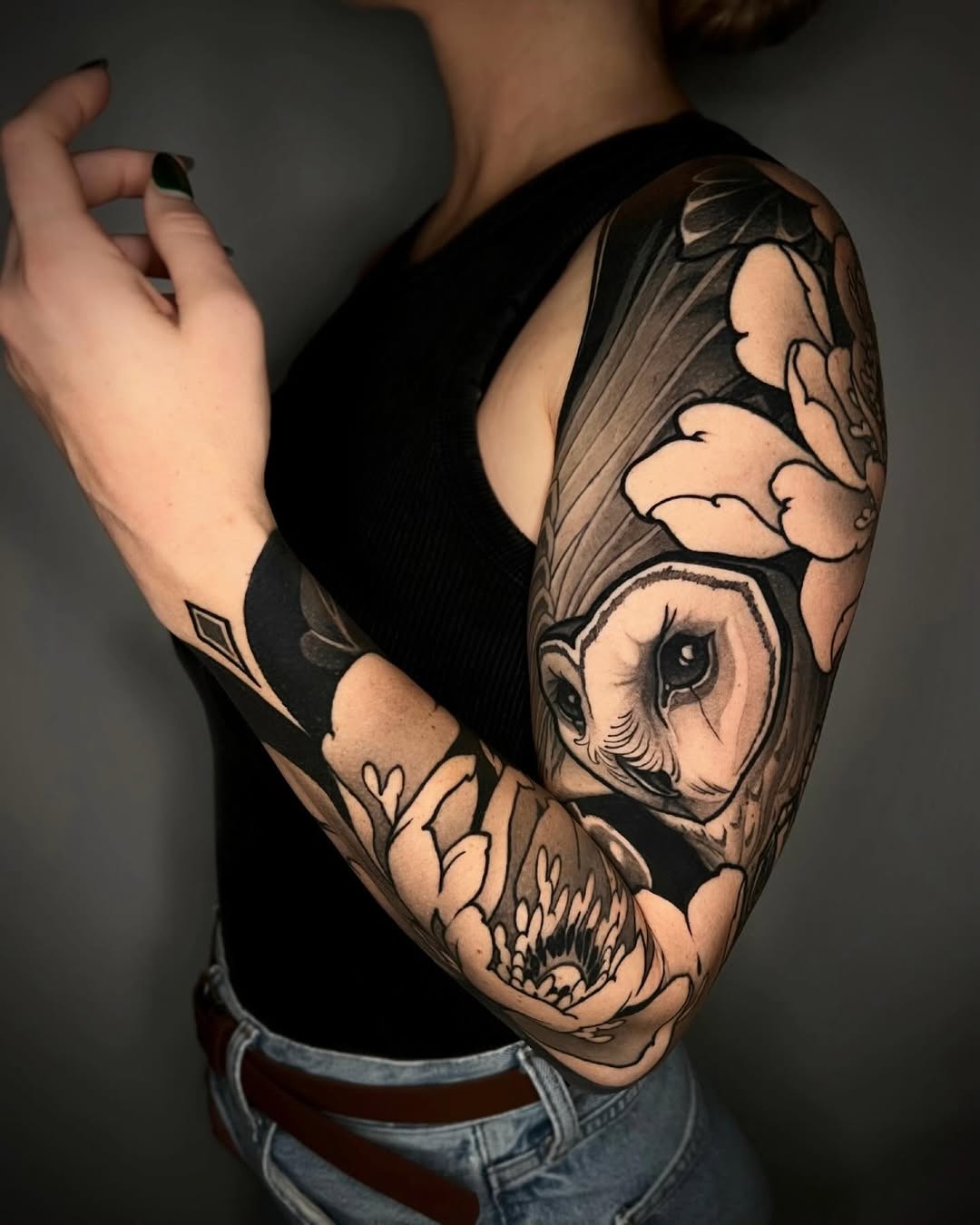 Black and grey sleeve tattoo featuring an owl with spread wings, skull, and botanical elements on the upper arm.